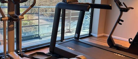Un gimnasio en casa con cinta de correr, elíptica y bicicleta estática, ventanas amplias con vistas a árboles y un estacionamiento.