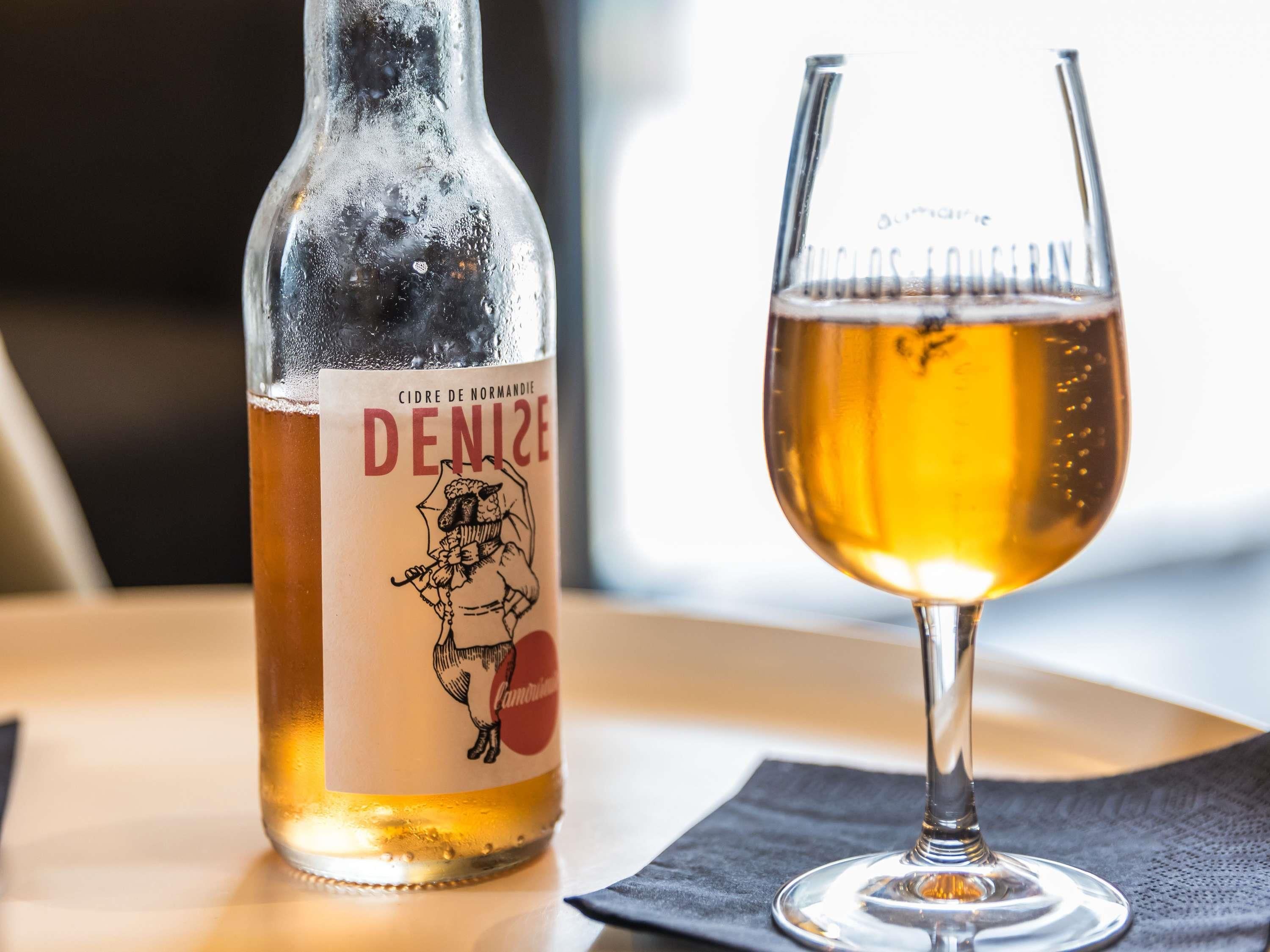 Una botella de Sidra de Normandía Denize con un vaso de líquido ámbar a su lado.
