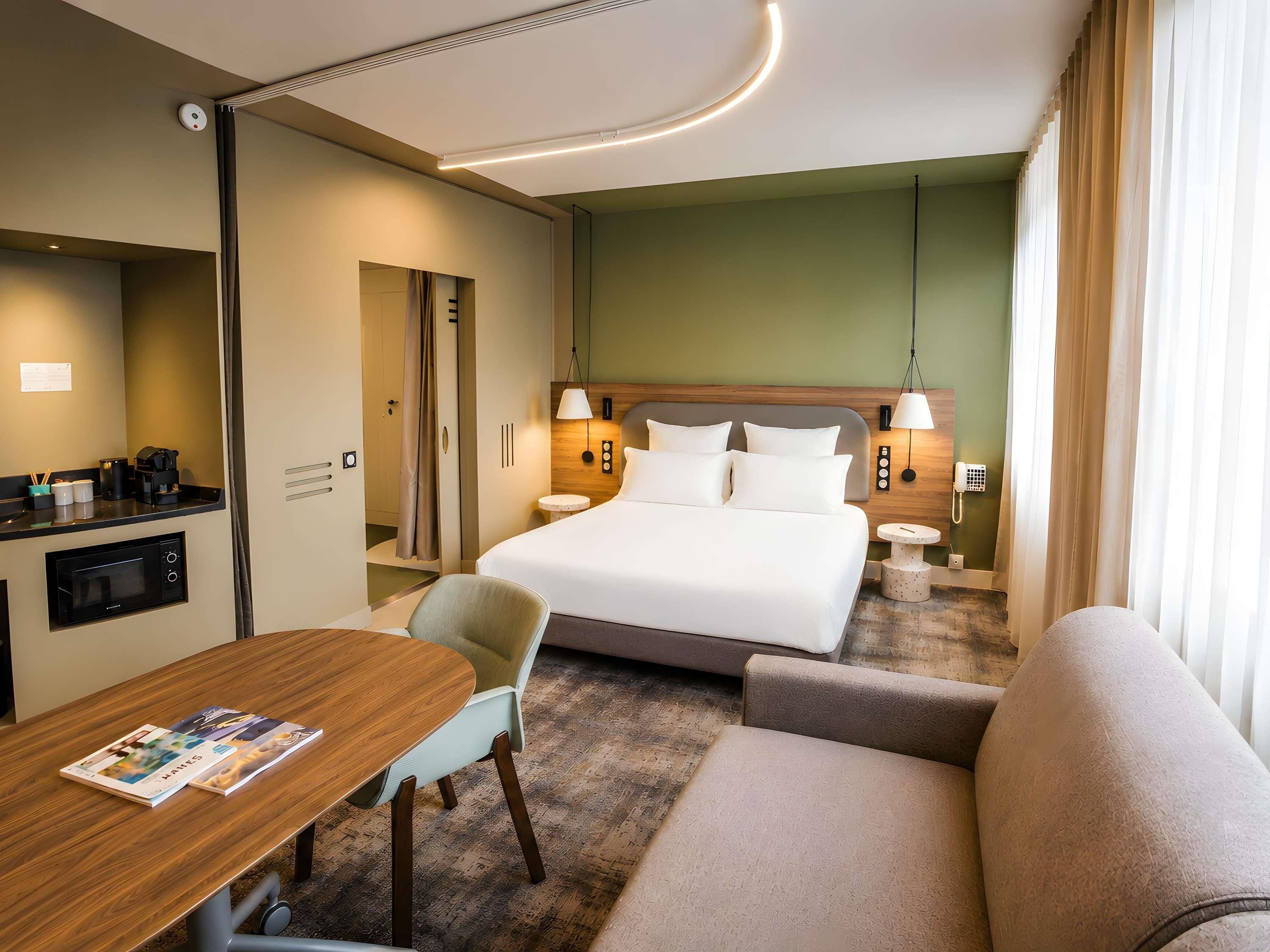 A hotel room with a bed, a desk, a chair, a sofa, a microwave, a coffee maker, a telephone, and a wall-mounted light fixture.