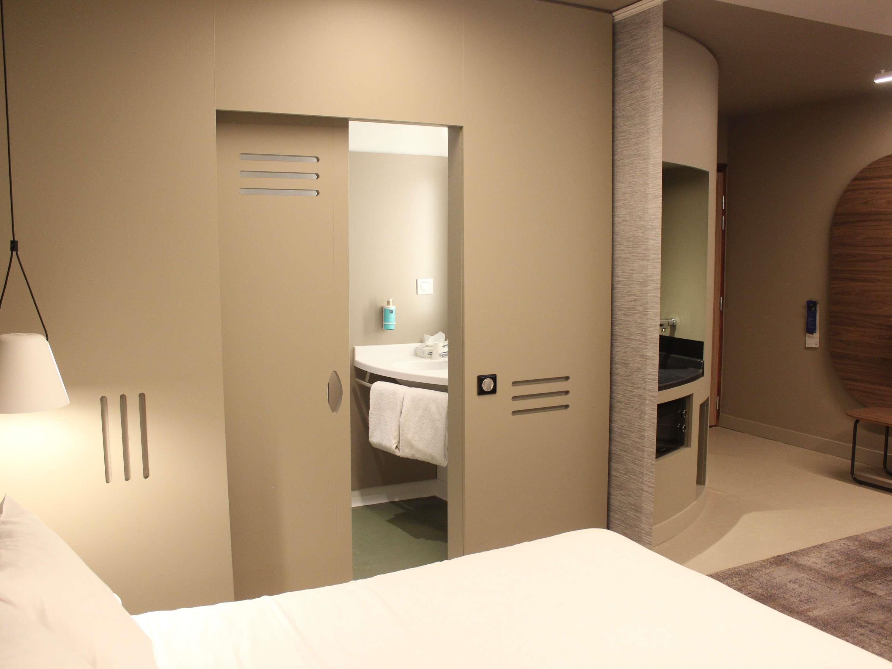 A modern hotel room with a bed, a bathroom with a sink and mirror, and a door leading to another room.