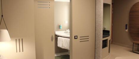 A modern hotel room with a bed, a bathroom with a sink and mirror, and a door leading to another room.
