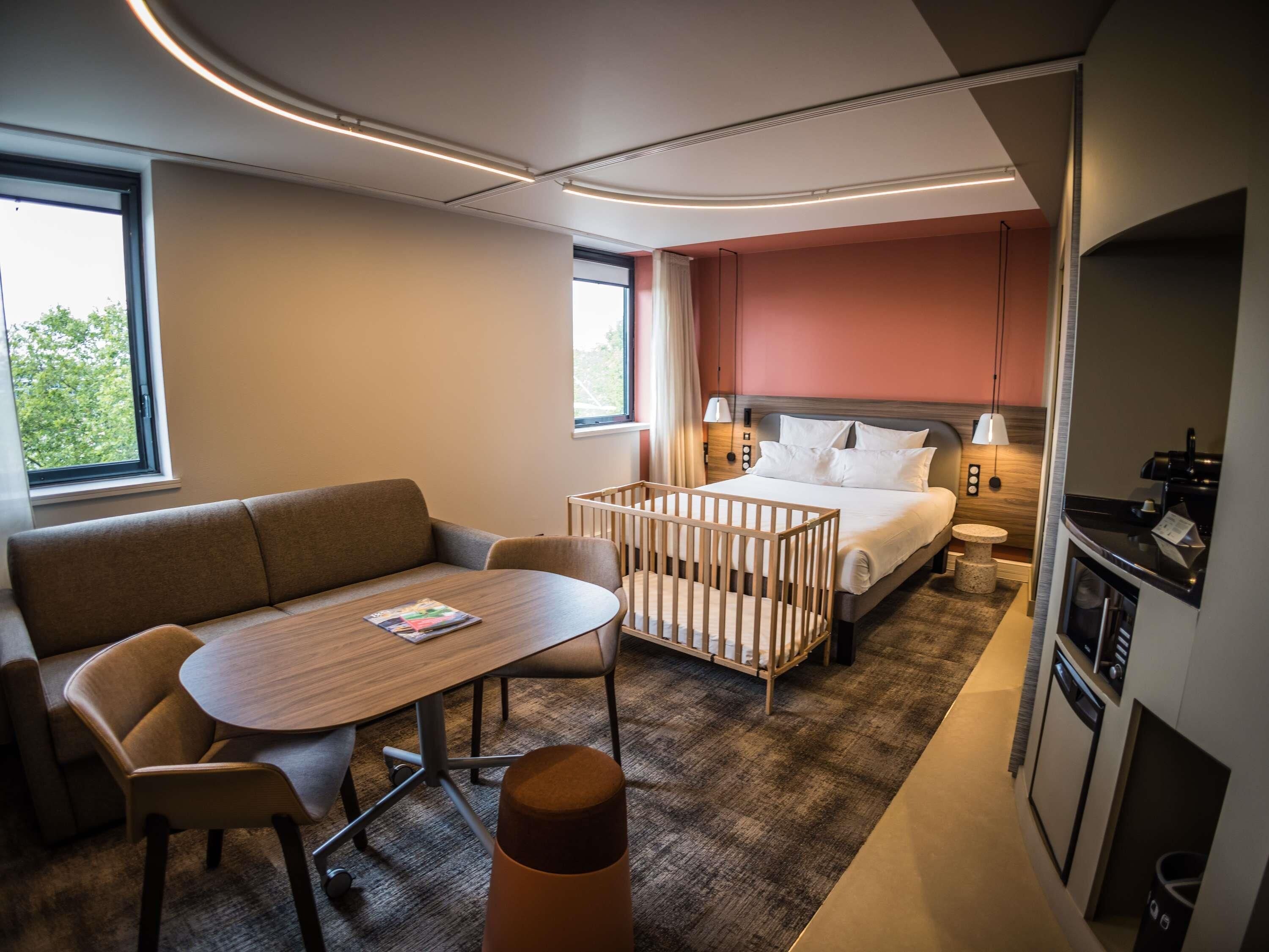 A modern hotel room with a bed, a seating area with a table, a crib, and a kitchenette.