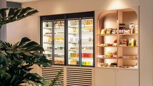 A refrigerated display case with various beverages and snacks, a wooden shelf with packaged goods, and a plant in the foreground.
