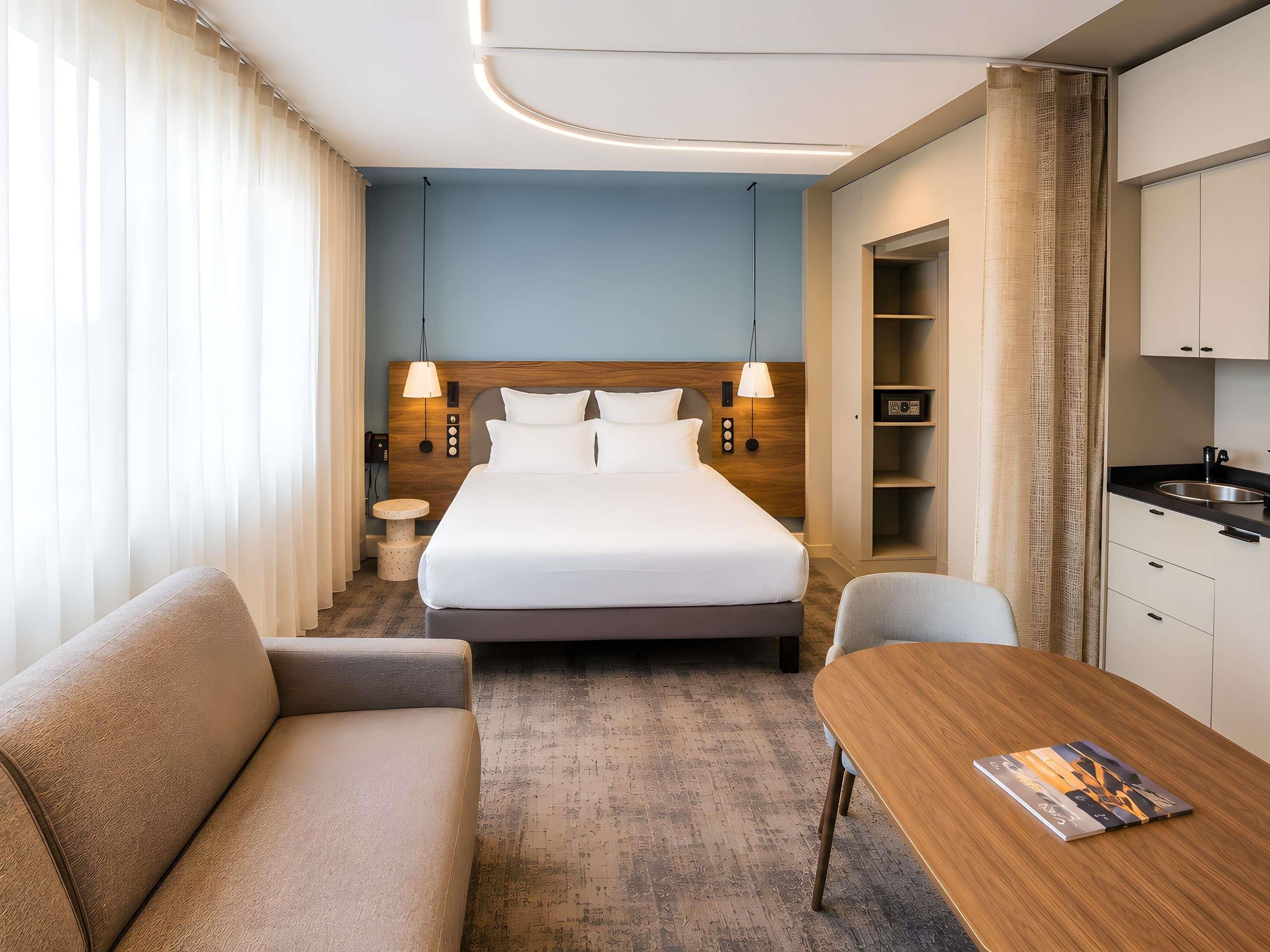 A hotel room with a bed, a sofa, a dining table, a small round table, and a kitchenette.