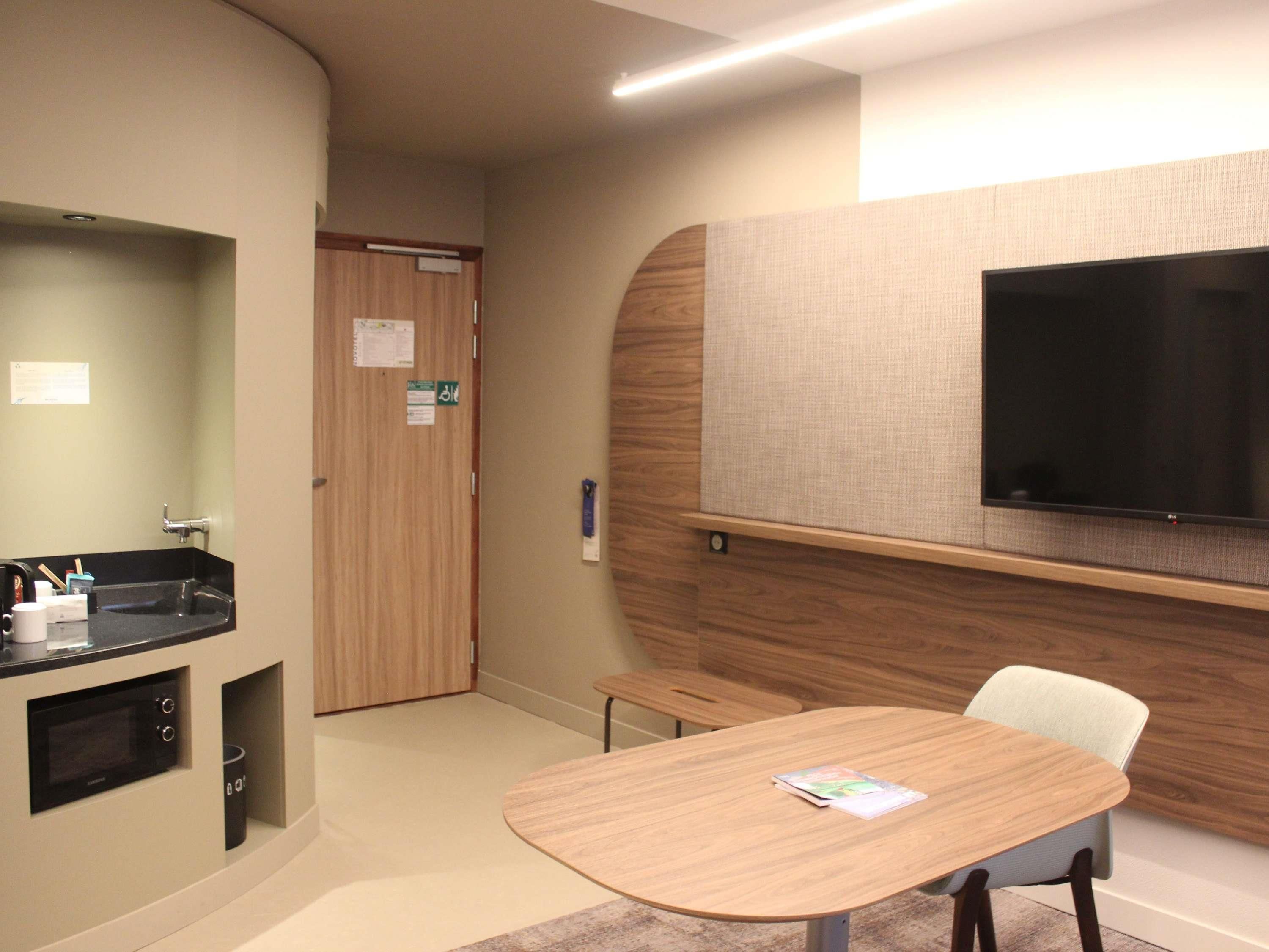 Habitación de hotel con kitchenette, televisor de pantalla plana montado en la pared, una mesa de madera y una sola silla.