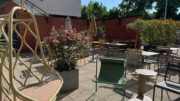 Un espacio al aire libre con sillas y mesas, una planta en maceta con flores rosas, y un edificio al fondo.