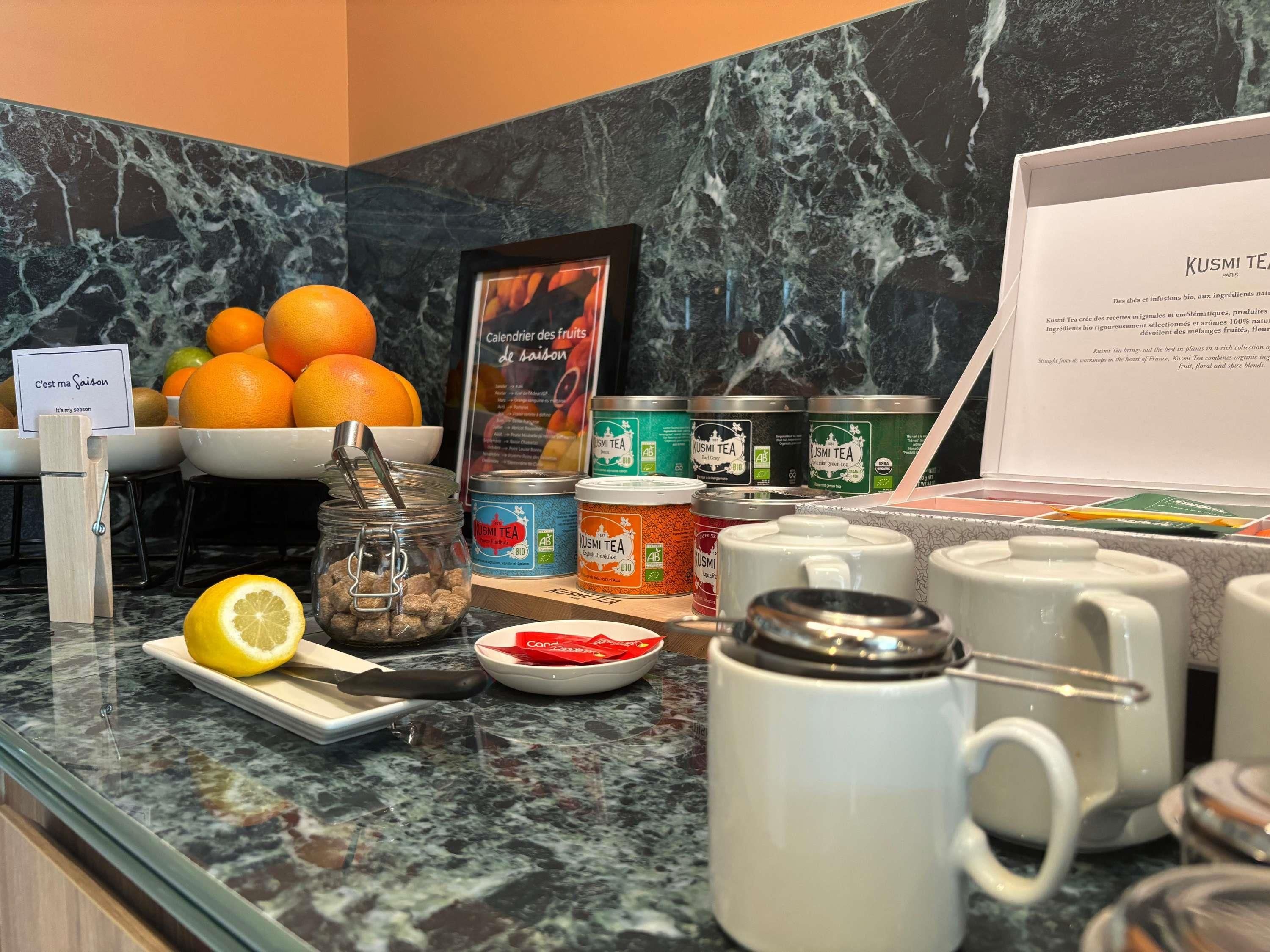 Uma mesa de café da manhã com uma tigela de laranjas, um limão, um pote de muesli e várias latas de chá Kusmi Tea.