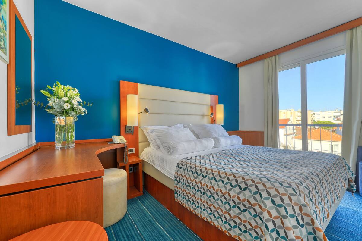 Ein Hotelzimmer mit einem großen Bett, einem Schreibtisch mit Telefon, einer Blumenvase und Blick auf Gebäude durch das Fenster.