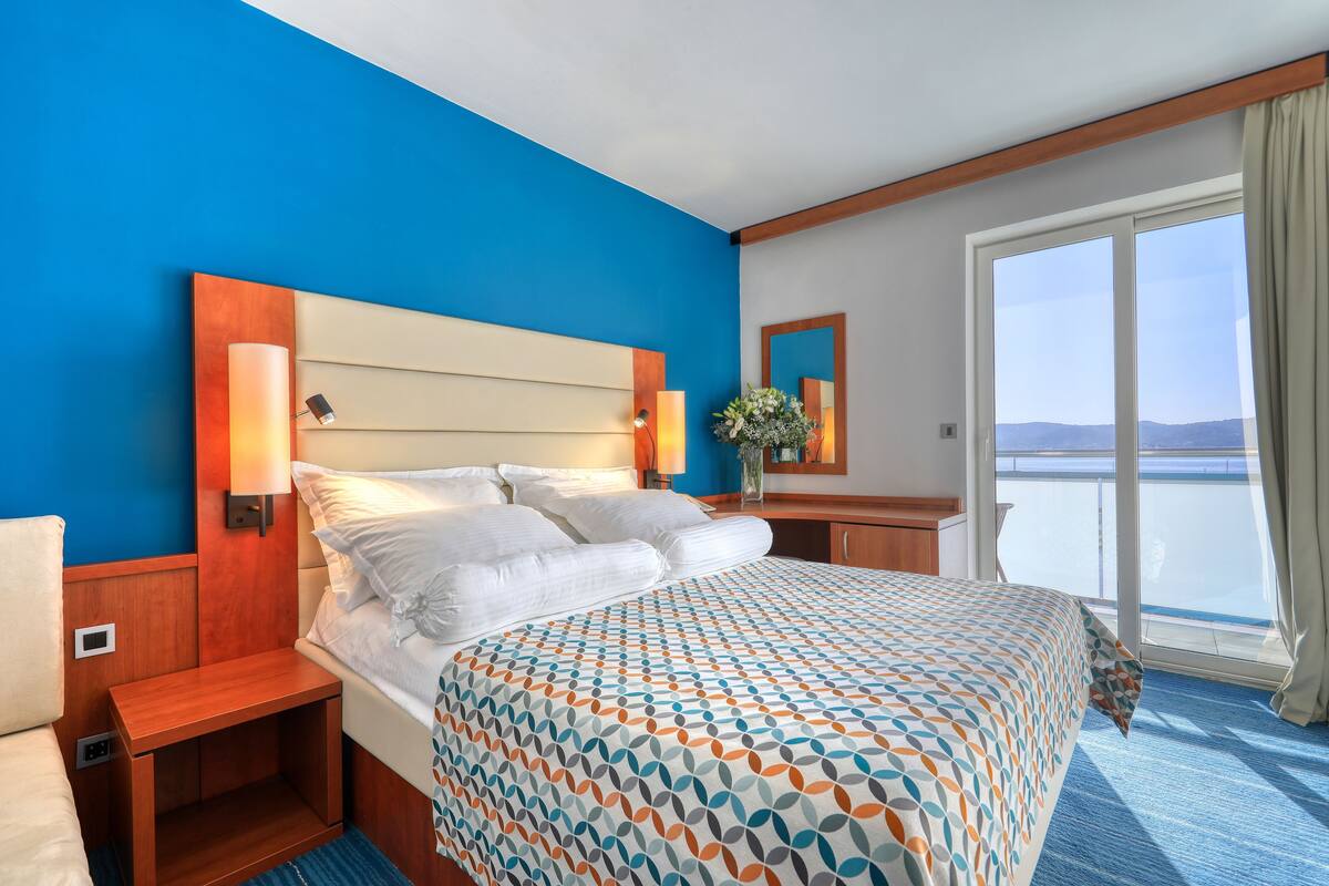 Ein Hotelzimmer mit einem großen Bett, einem Nachttisch, einer Lampe, einem Spiegel und Blick auf das Meer durch ein großes Fenster.