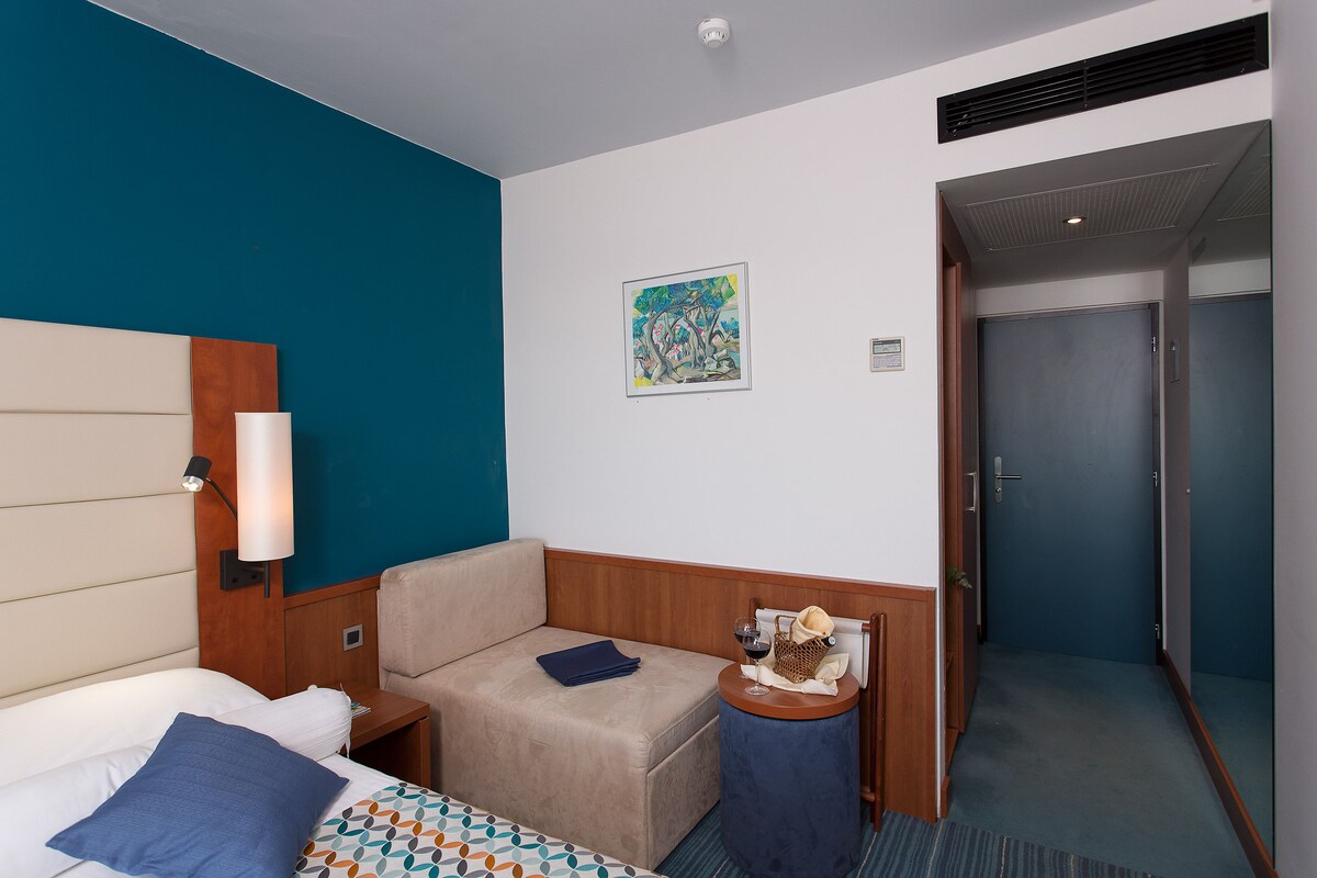 Une chambre d’hôtel avec un lit, un canapé, une petite table avec une lampe, un tableau au mur et une porte donnant sur une autre pièce.