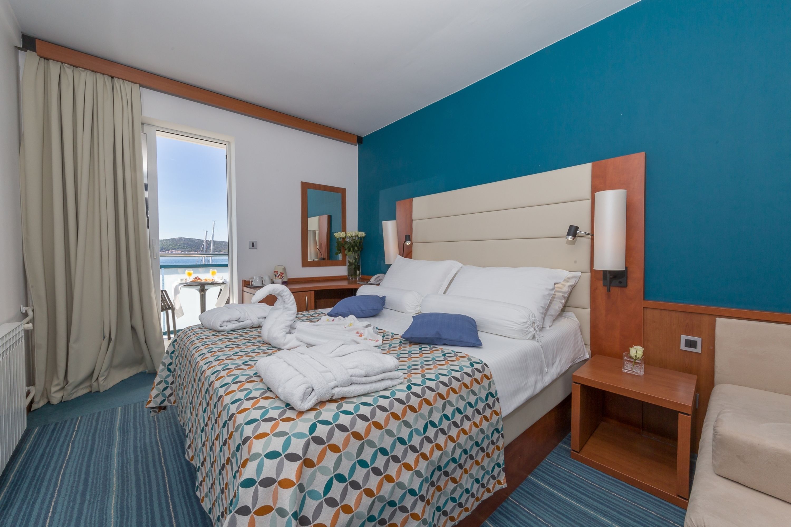 A hotel room with a large bed, a nightstand, a chair, a view of the sea, and a window with curtains.