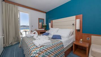 A hotel room with a large bed, a nightstand, a chair, a view of the sea, and a window with curtains.