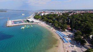 An aerial view of a coastal area with a beach, clear blue waters, and a row of beach umbrellas. - Hotel Kornati Biograd (Biograd na Moru)