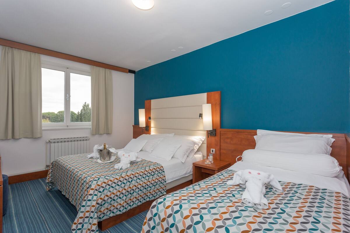 Ein Hotelzimmer mit zwei Betten, einem Fenster mit Vorhängen, einem Holz-Kopfteil und einer blauen Akzentwand.