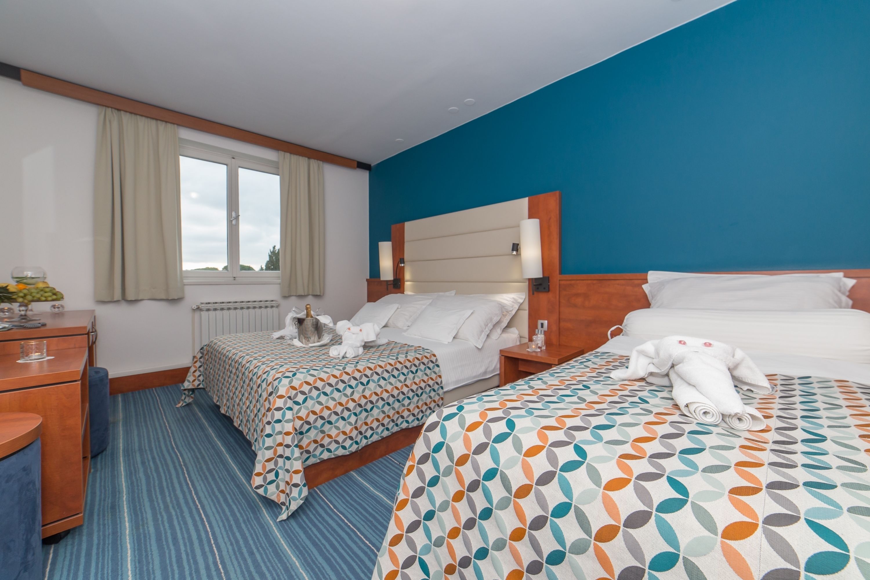 A hotel room with two beds, a wooden dresser, a window with curtains, and a view of the outdoors.