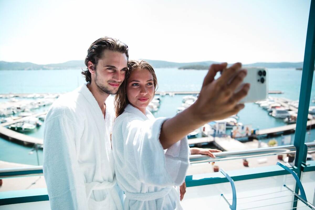 Ein Paar in weißen Gewändern macht auf dem Balkon ein Selfie mit Blick auf eine Marina.
