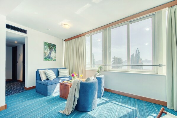 A hotel room with a blue carpet, a sofa, ottomans, a coffee table, and a large window with curtains.