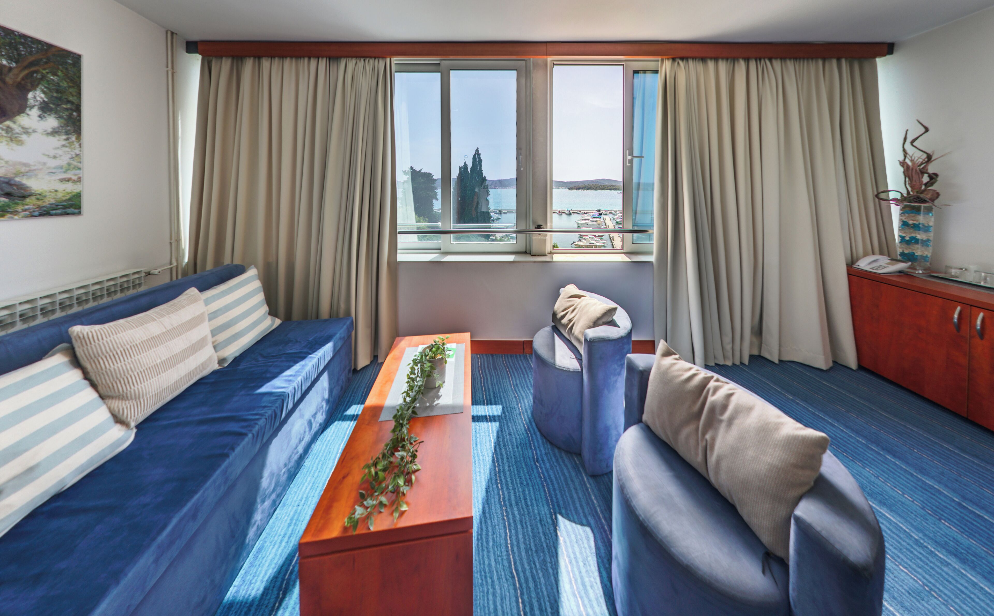 Habitació d'hotel amb un sofà blau, una taula auxiliar de fusta amb una planta, un petit seient i vista a una massa d'aigua a través de grans finestres.