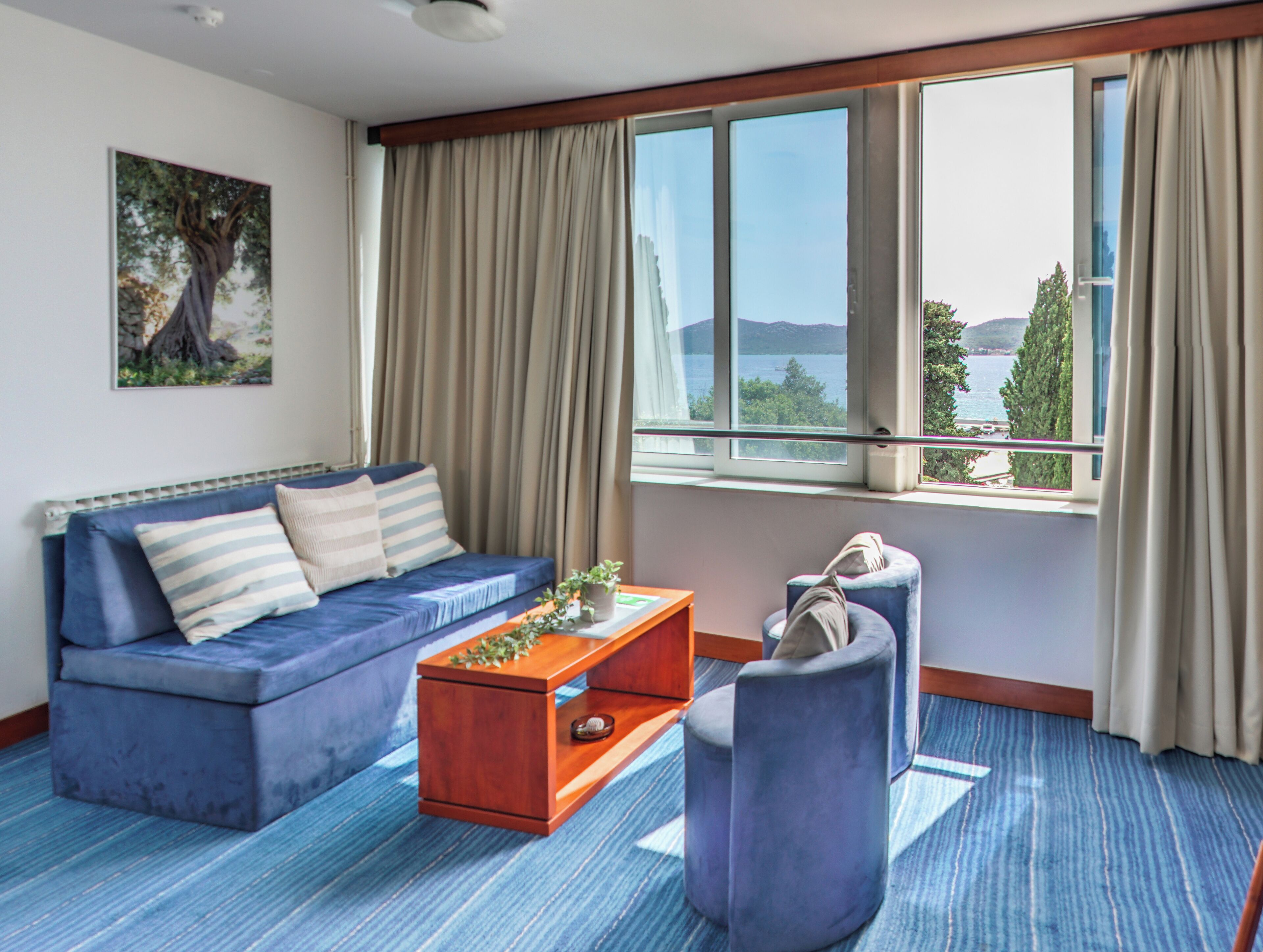 Une chambre d’hôtel moderne avec un canapé bleu, une table basse en bois, un fauteuil et une grande fenêtre offrant une vue sur la mer et les montagnes.