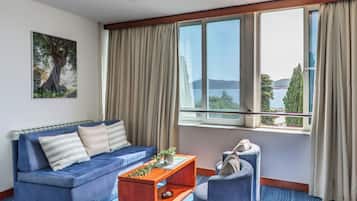 Une chambre d’hôtel moderne avec un canapé bleu, une table basse en bois, un fauteuil et une grande fenêtre offrant une vue sur la mer et les montagnes.