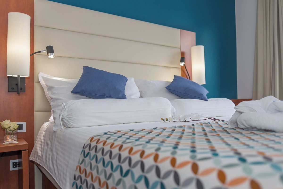 Un lit bien fait, avec des draps blancs et des oreillers bleus, une couverture à motifs et des lampes de chevet.