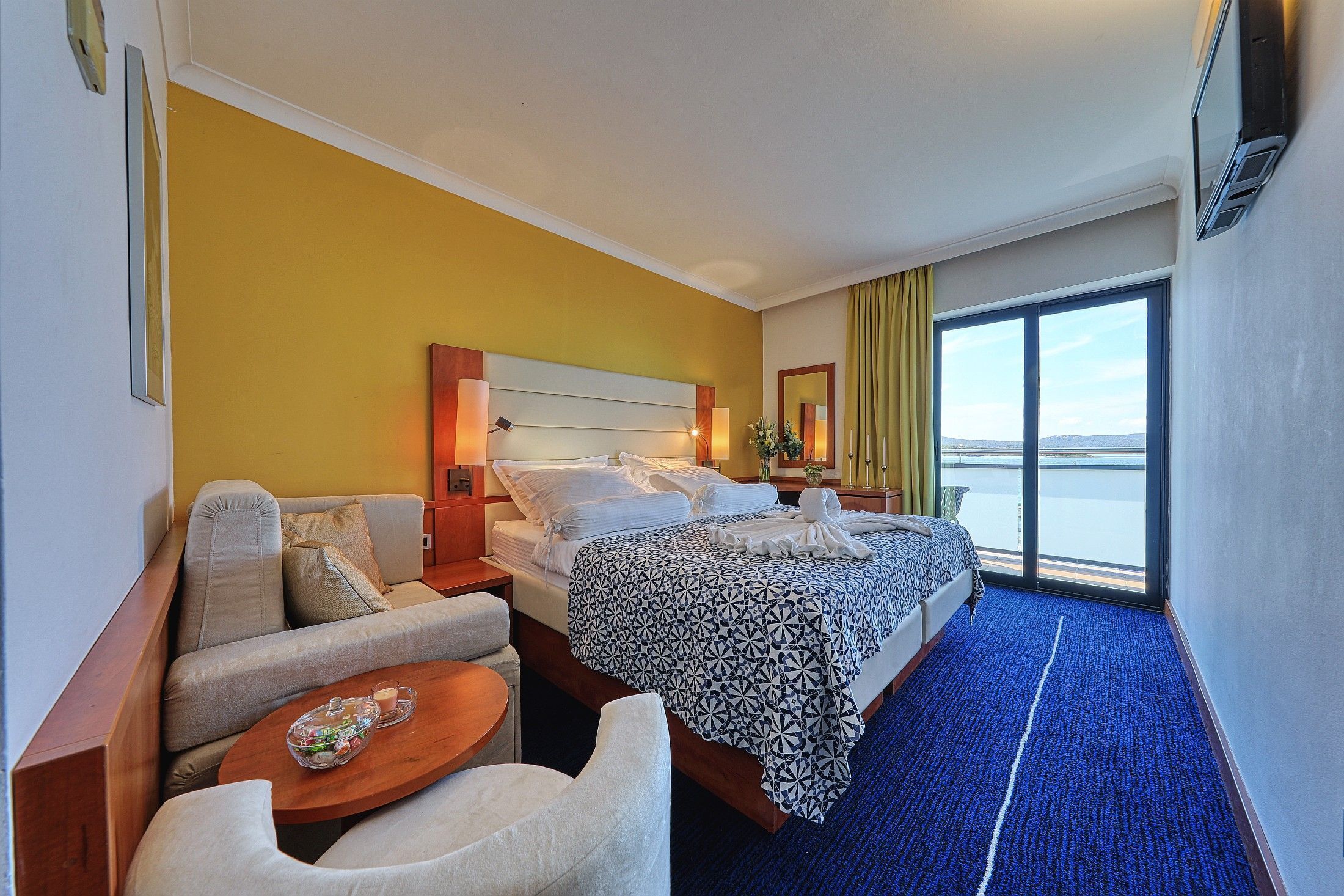 A hotel room with a bed, a sofa, a small table, and a view of the sea through large windows.