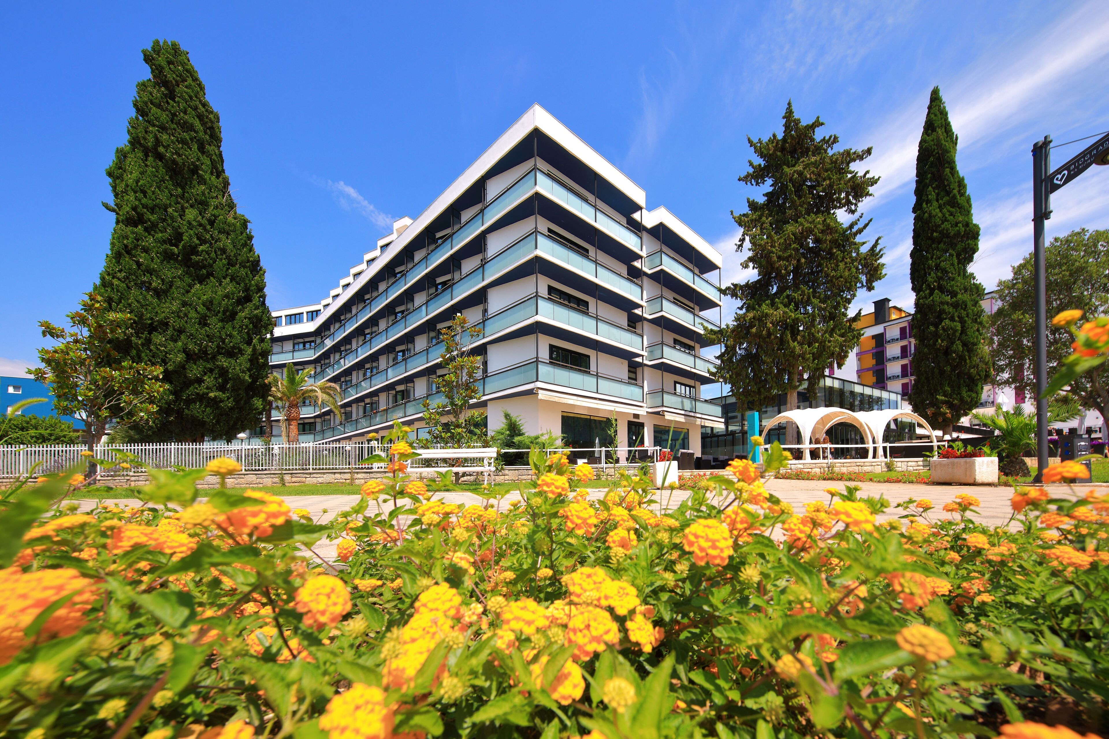 A modern multi-story building with balconies, surrounded by a landscaped area with flowering plants.