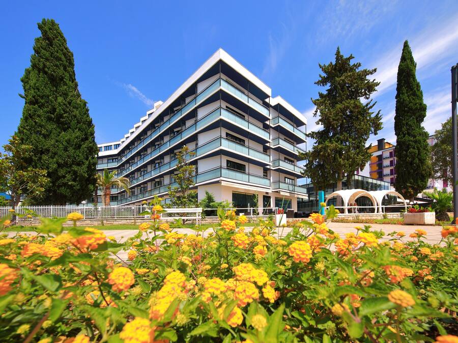A modern multi-story building with balconies, surrounded by a landscaped area with flowering plants.