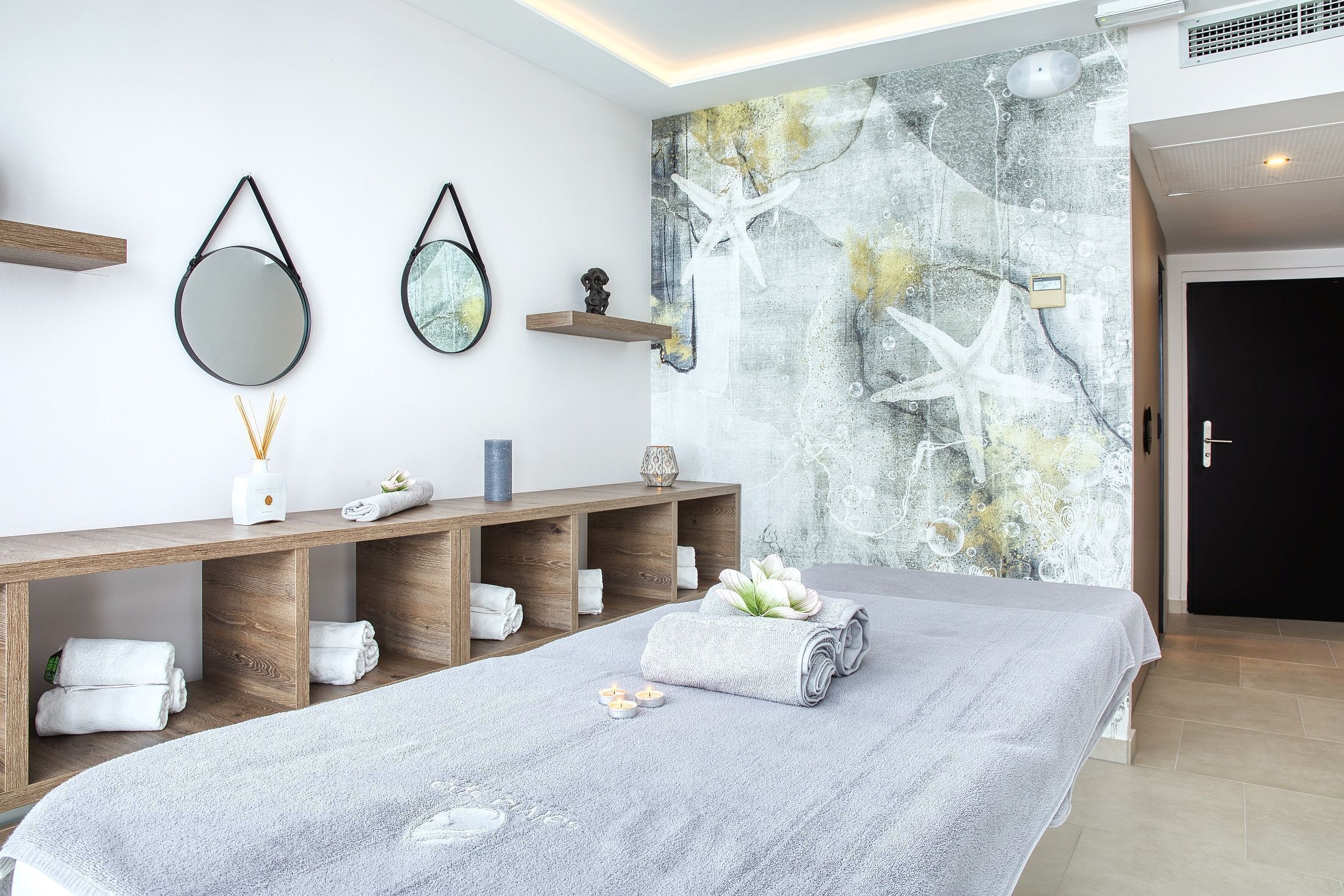 Um spa moderno com uma cama grande, prateleiras de madeira com toalhas, um espelho fixado na parede e um mural abstrato decorativo.