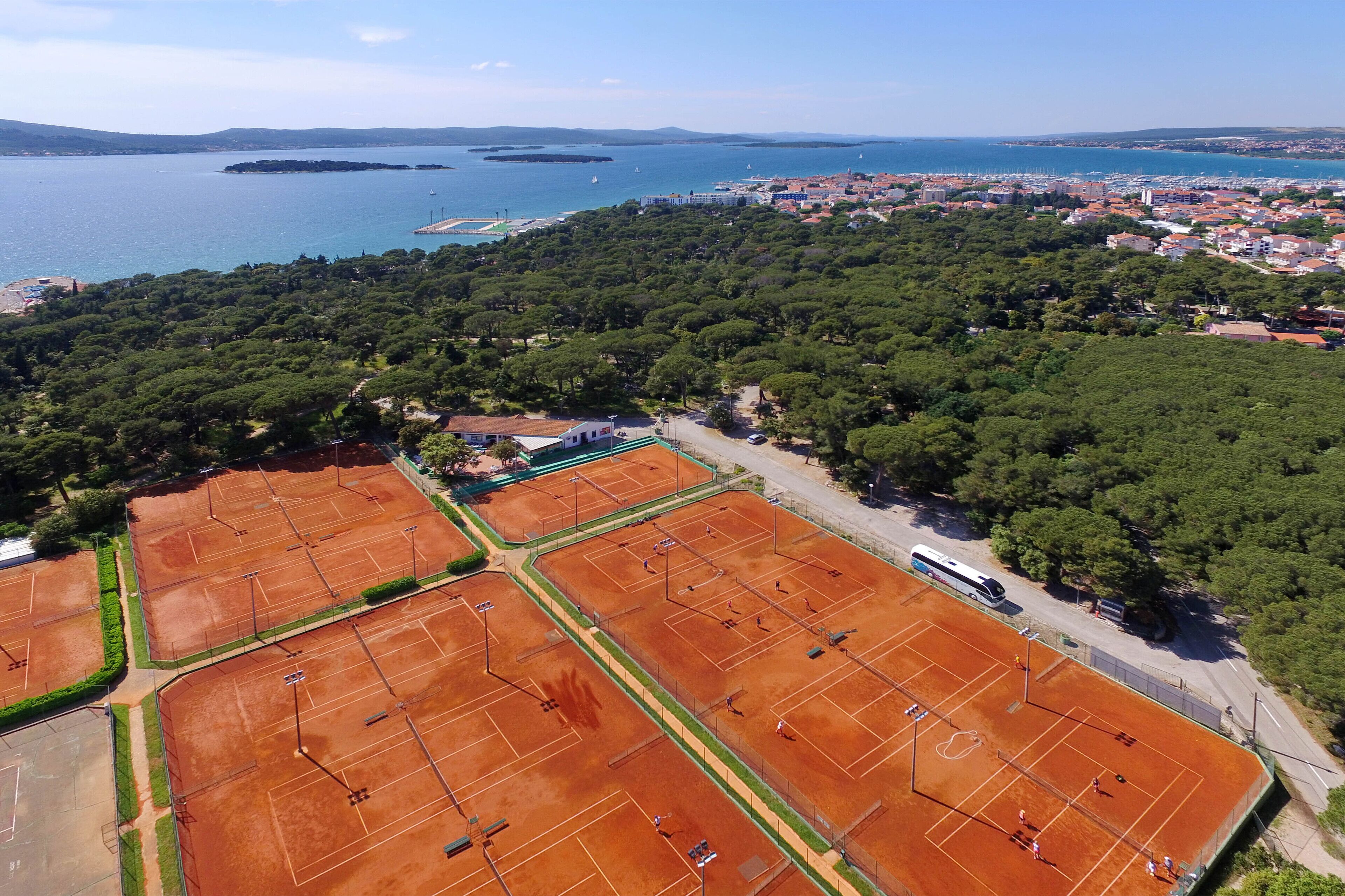 An aerial view of a coastal area with tennis courts, a bus, and a cityscape in the background.