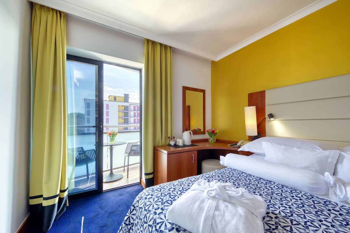 Une chambre d’hôtel comprenant un lit, un bureau, une chaise, une petite table avec un vase de fleurs et donnant sur le balcon extérieur.
