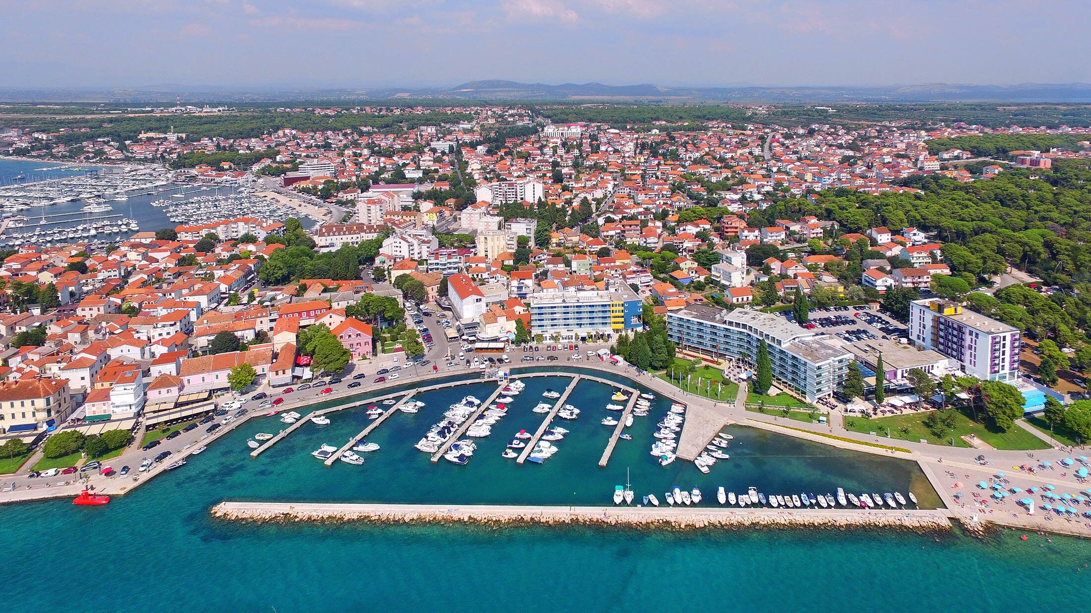 Vista aerea di una cittadina costiera con un porto turistico, edifici residenziali e un grande hotel con diversi piani e balconi.