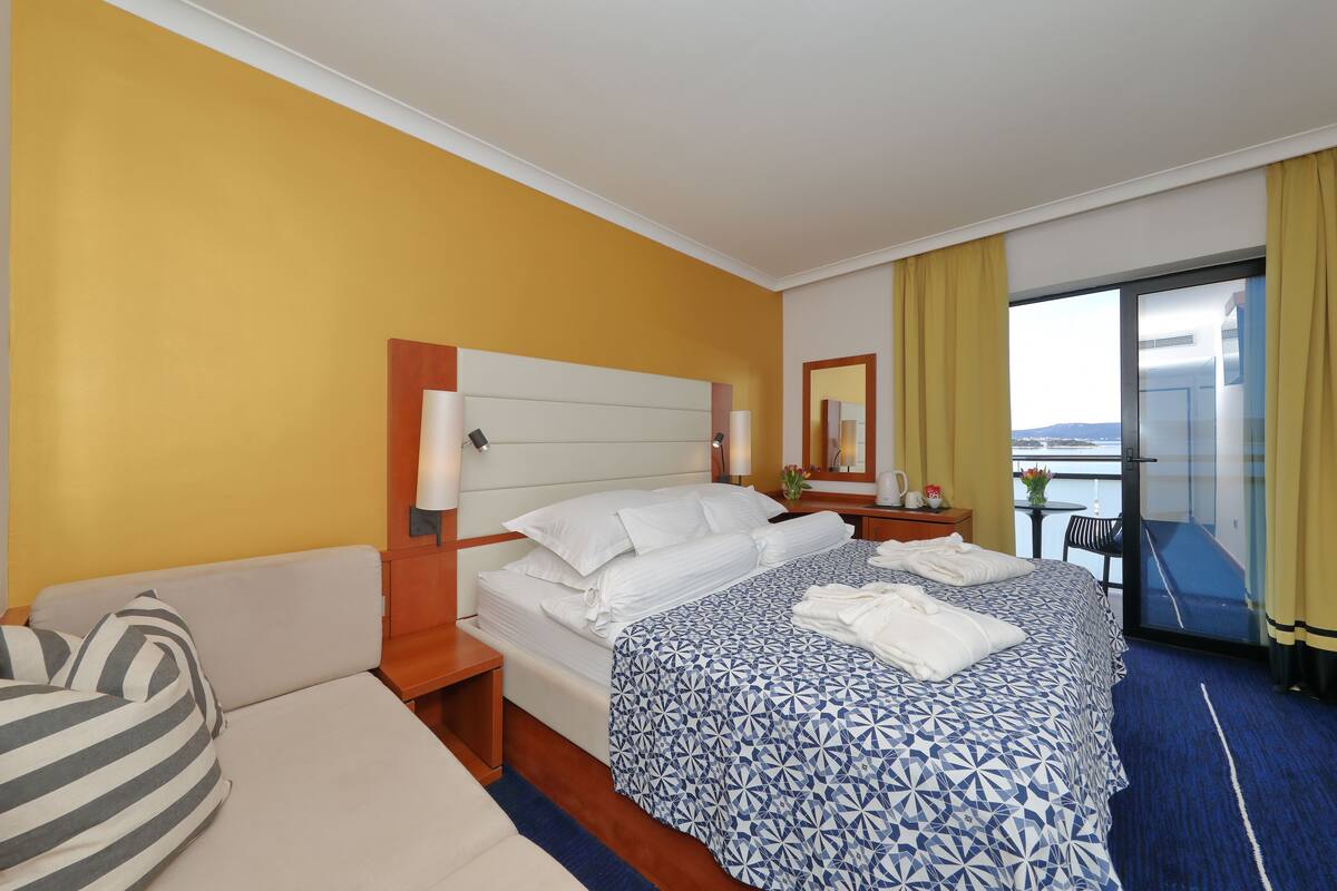 Une chambre d’hôtel comprenant un lit, un canapé, une table de chevet, une table avec une chaise, et donnant sur l’extérieur.