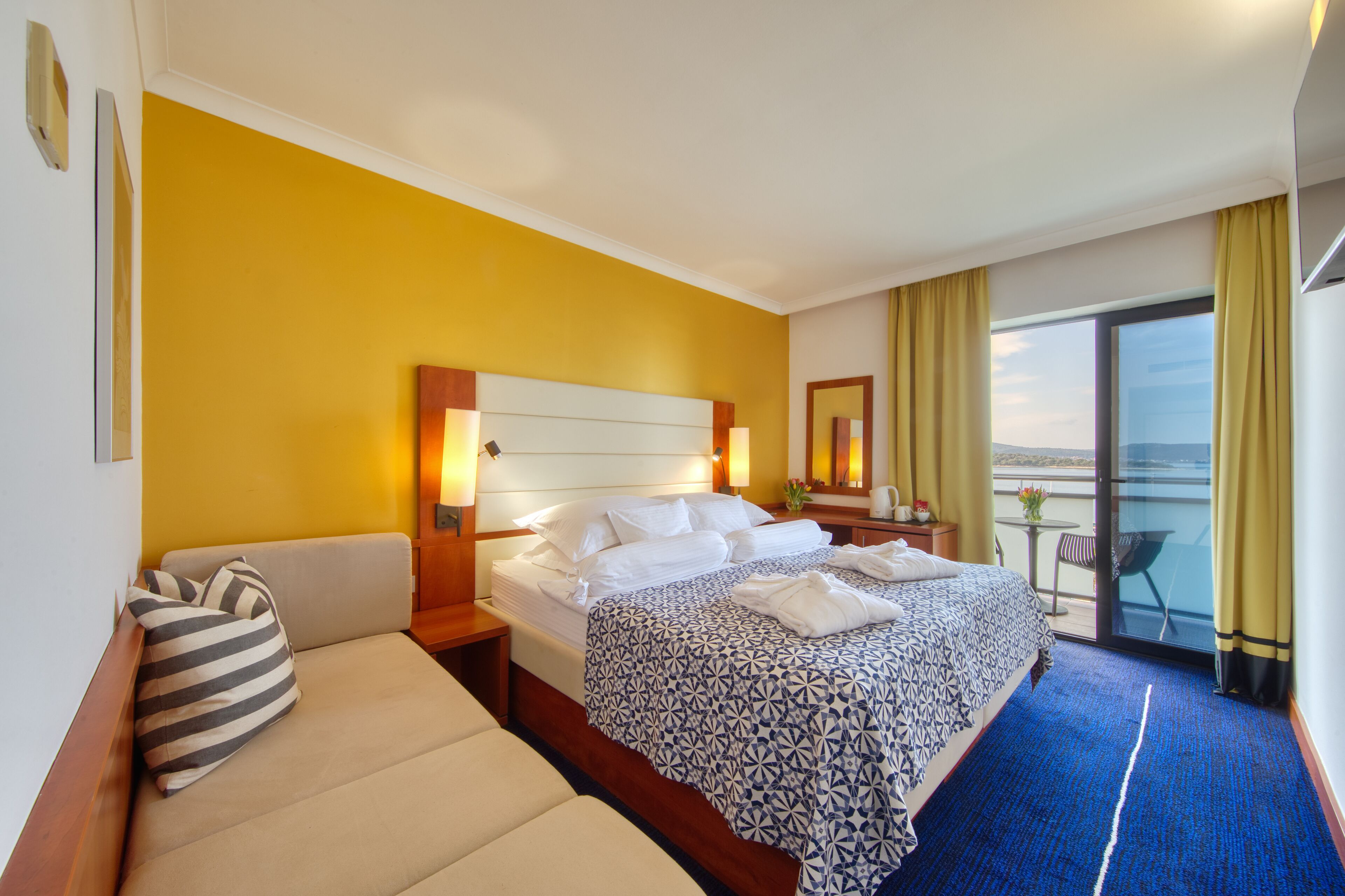 A hotel room with a bed, a sofa, a small table with a chair, and a balcony with a view.