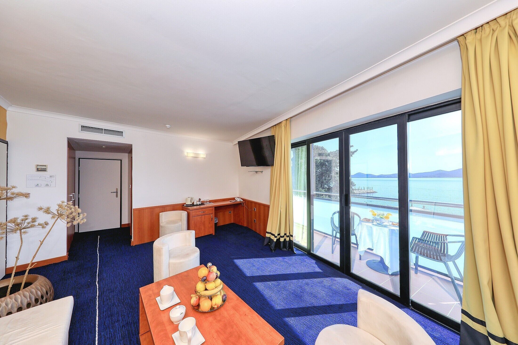 A hotel room with a balcony, a dining table with fruit, a desk, and a TV.