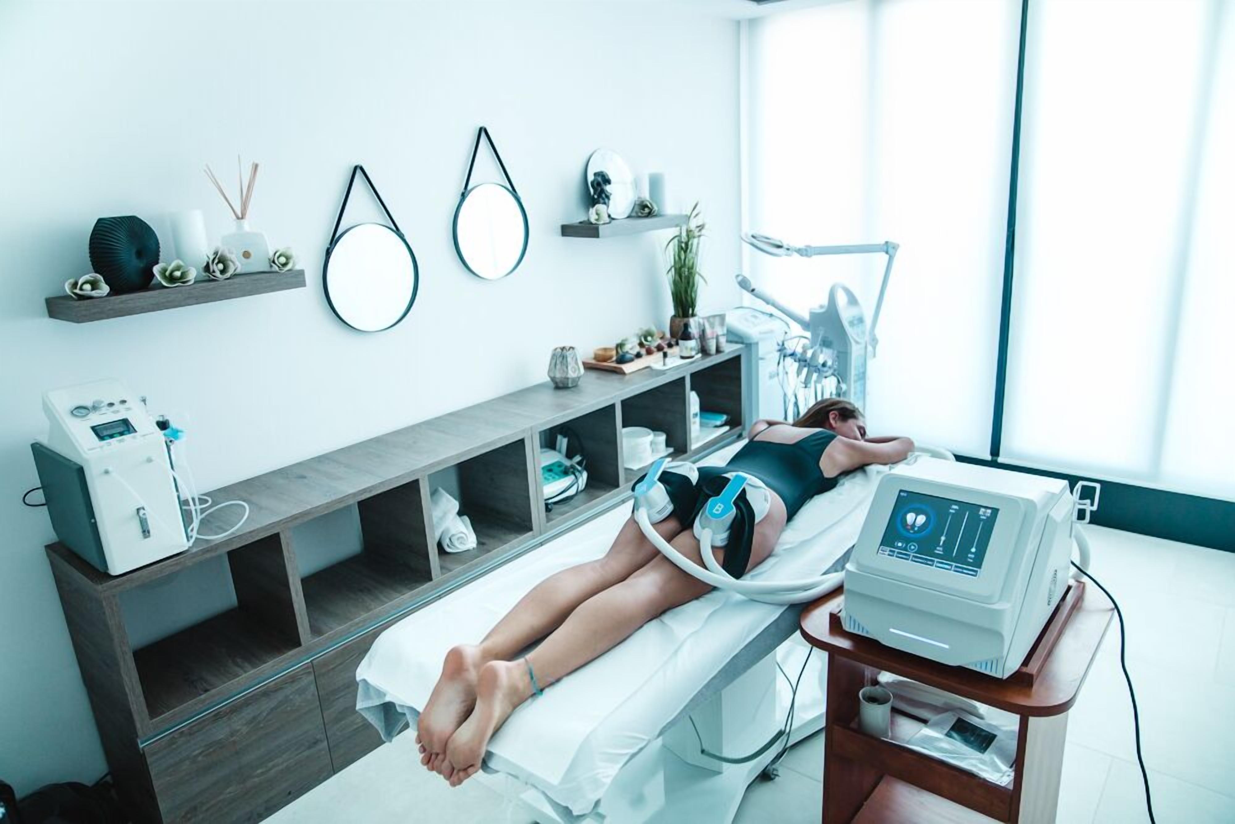 A medical examination room with a patient lying on a bed, a medical device on a cart, and shelves with various items.
