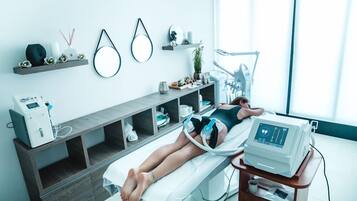 A medical examination room with a patient lying on a bed, a medical device on a cart, and shelves with various items.