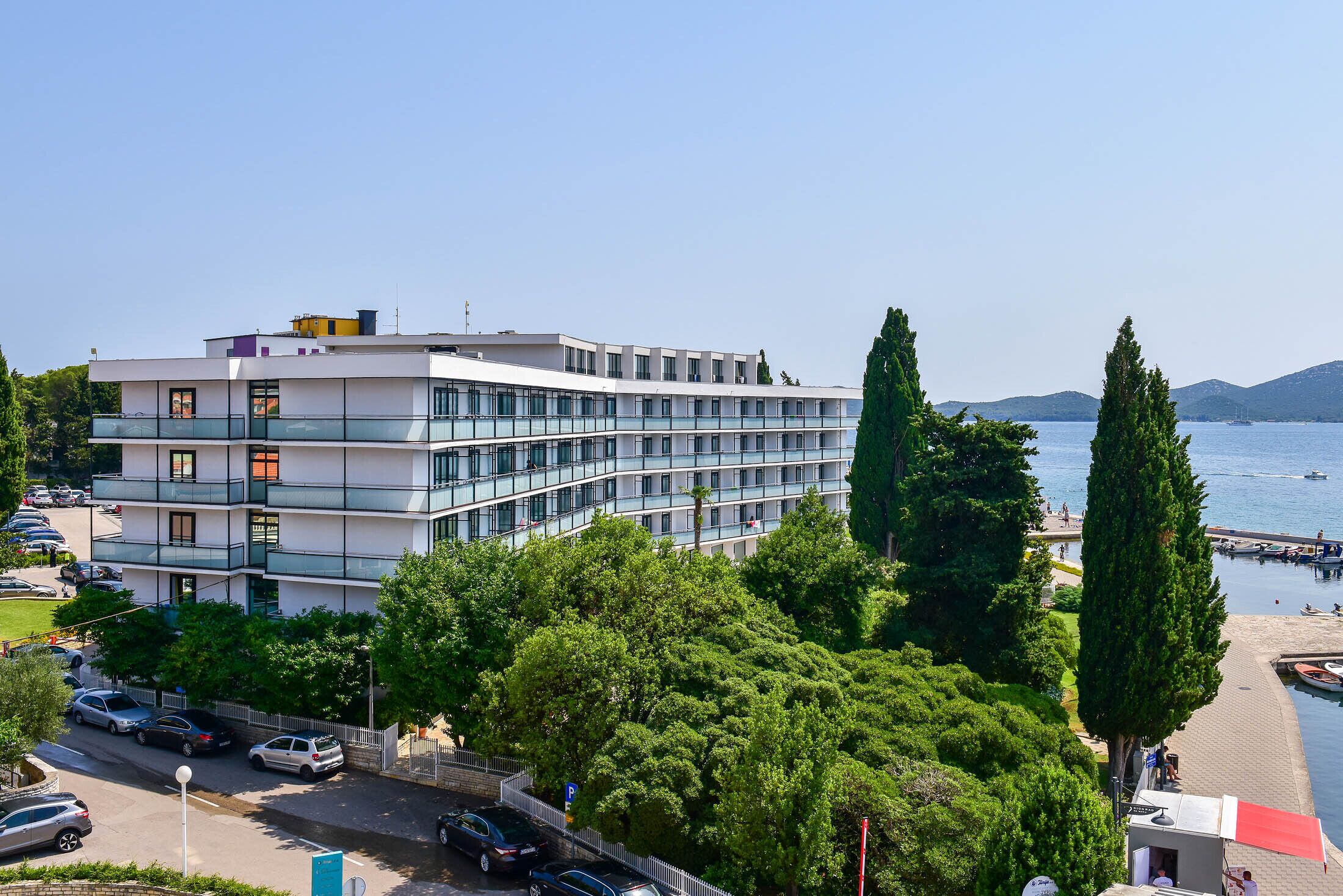 A multi-story building with a modern design, located near a waterfront with trees and parked cars.