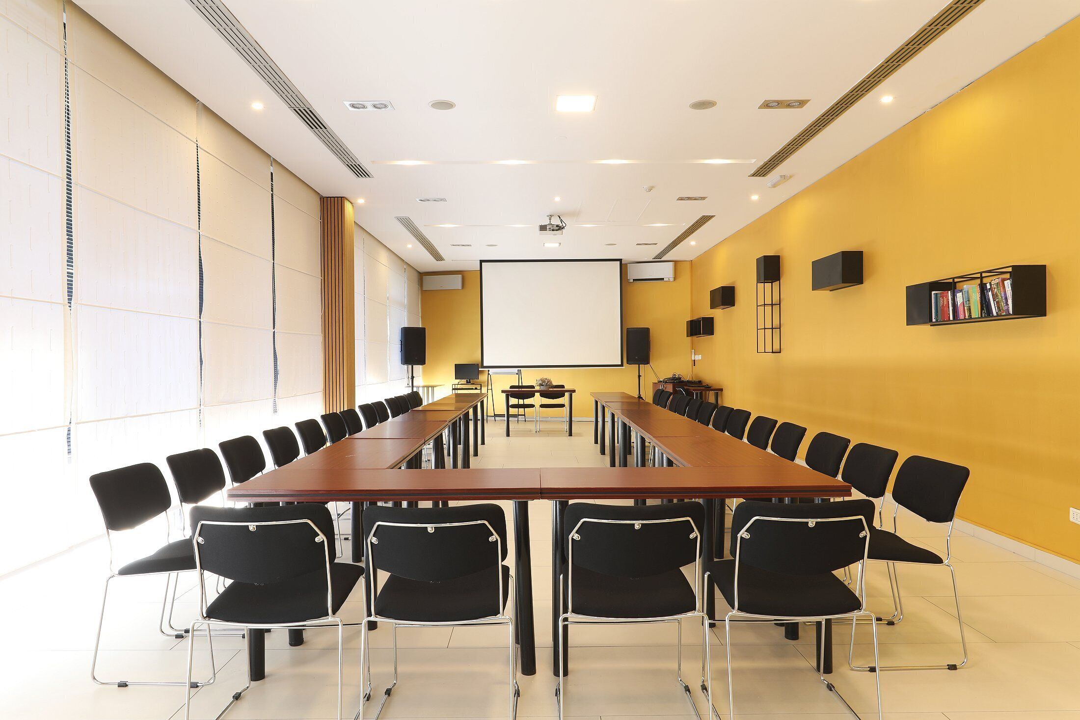 A conference room with a long table, black chairs, a projector screen, and a bookshelf.