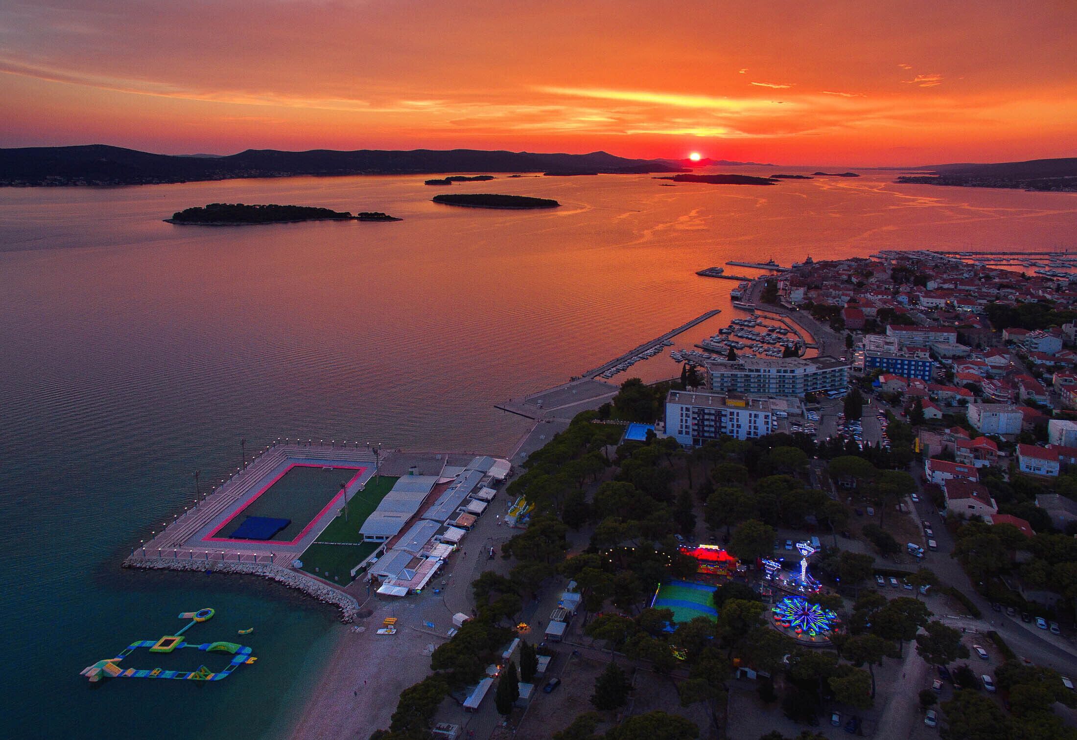 Luchtbeeld van een kuststad bij zonsondergang, met een jachthaven, woongebouwen en een sportcomplex.