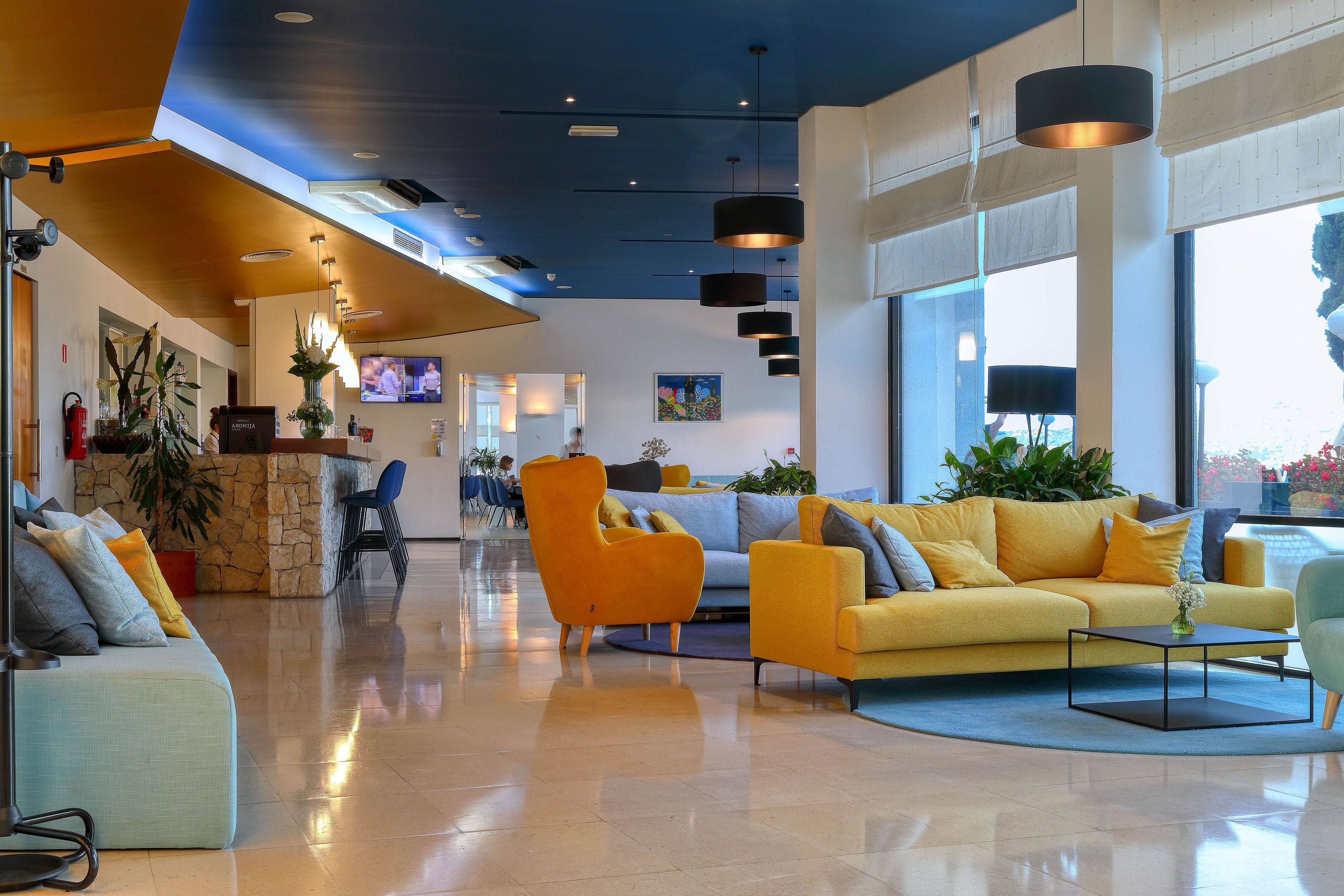 A modern lobby with a yellow sofa, orange armchair, and a blue couch. There are large windows, a TV, and a reception desk with a plant.