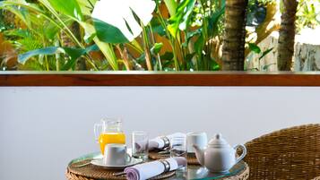 A wicker table set with a pitcher, glasses, a rolled napkin, a teapot, and a cup, with a view of lush greenery and palm trees in the background.