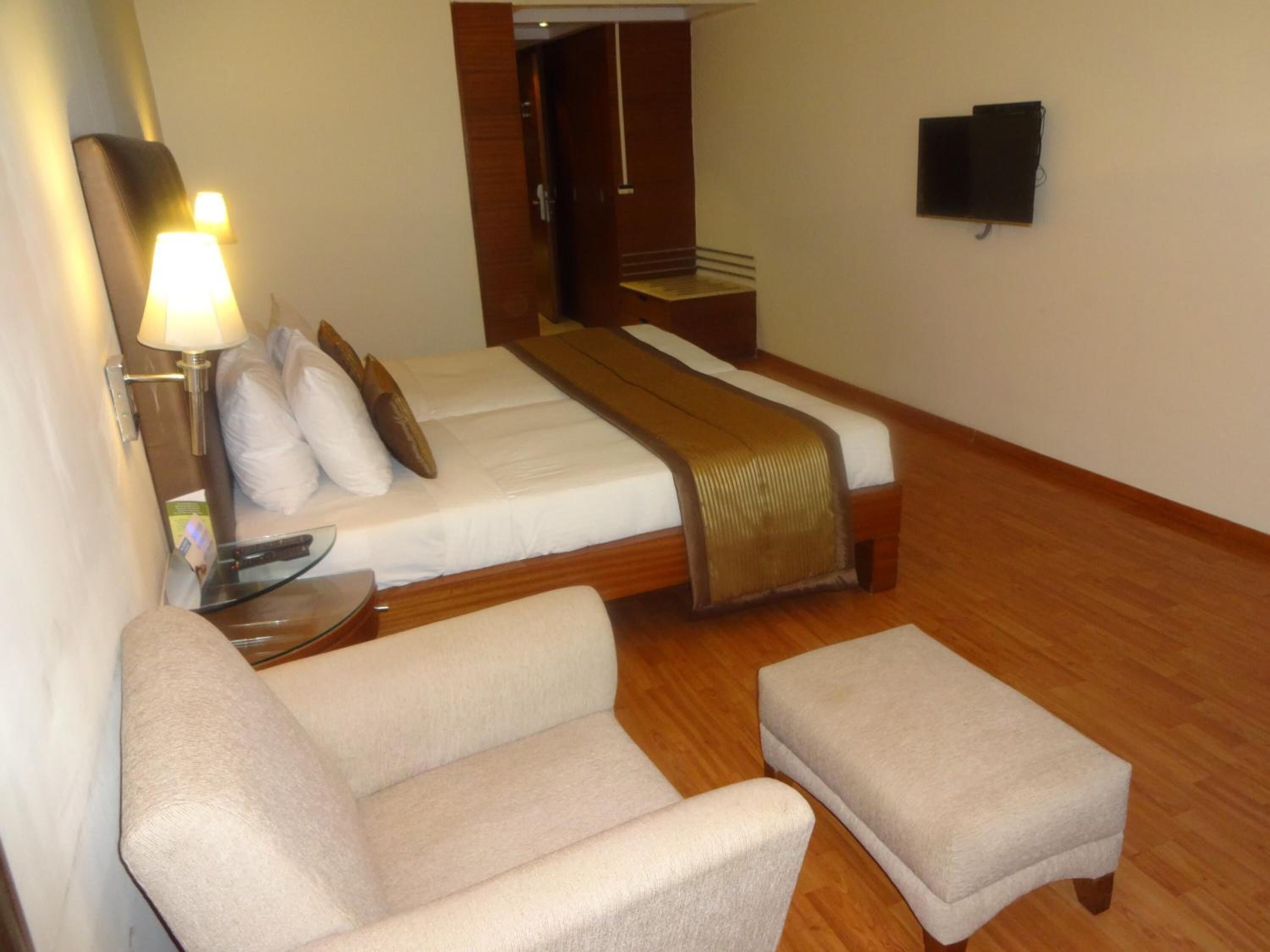 A hotel room with two beds, a sofa, an ottoman, a bedside table, a lamp, and a TV.