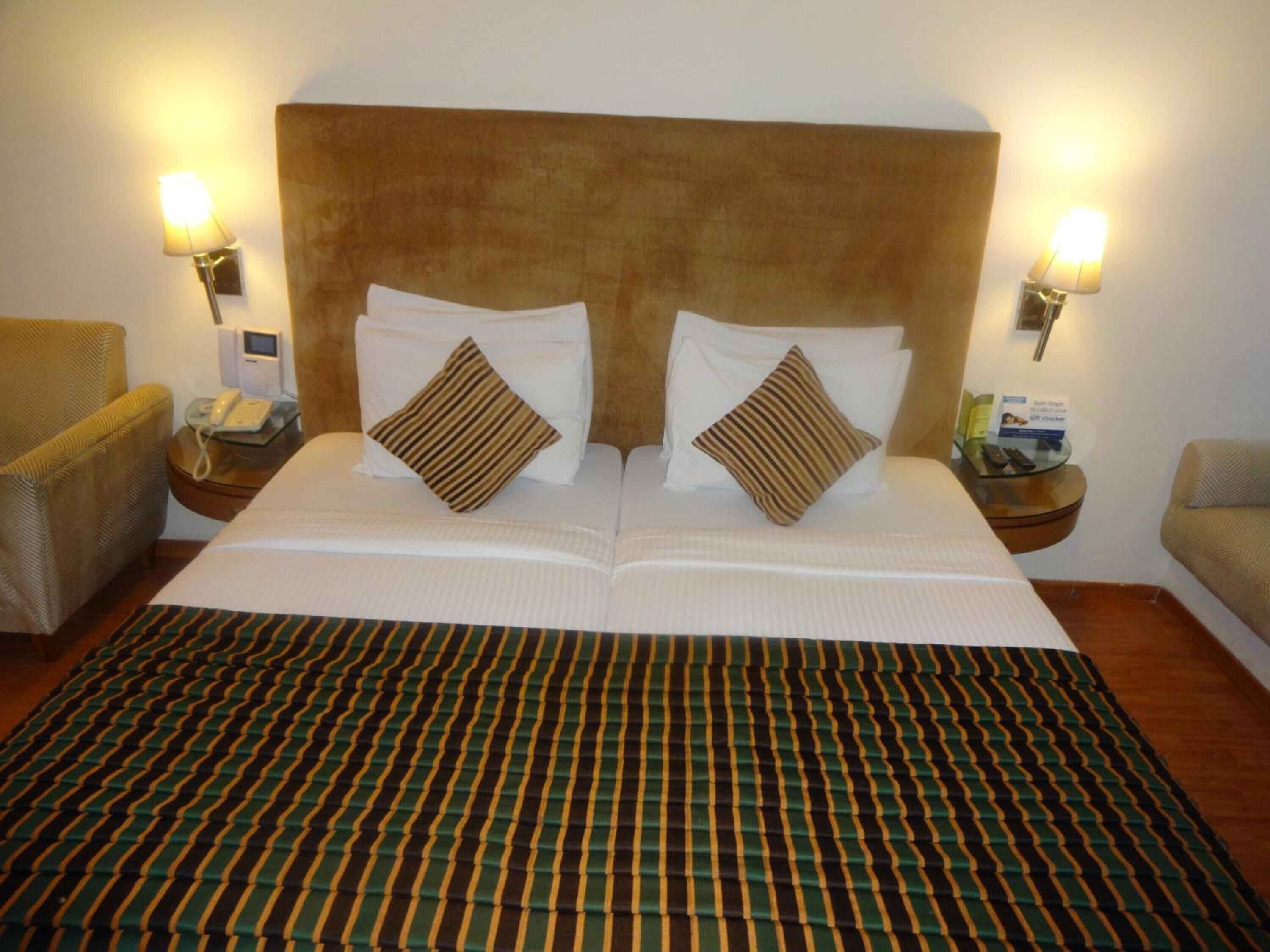 A double bed with striped bedding, two pillows, bedside lamps, a telephone, and a small table with a brochure.