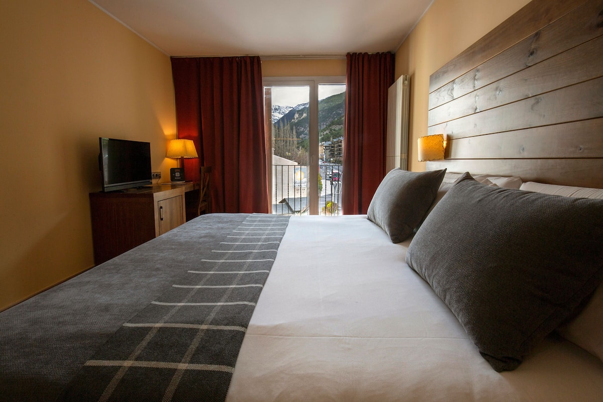 A hotel room with a bed, a TV, a lamp, and a view of mountains through the window.