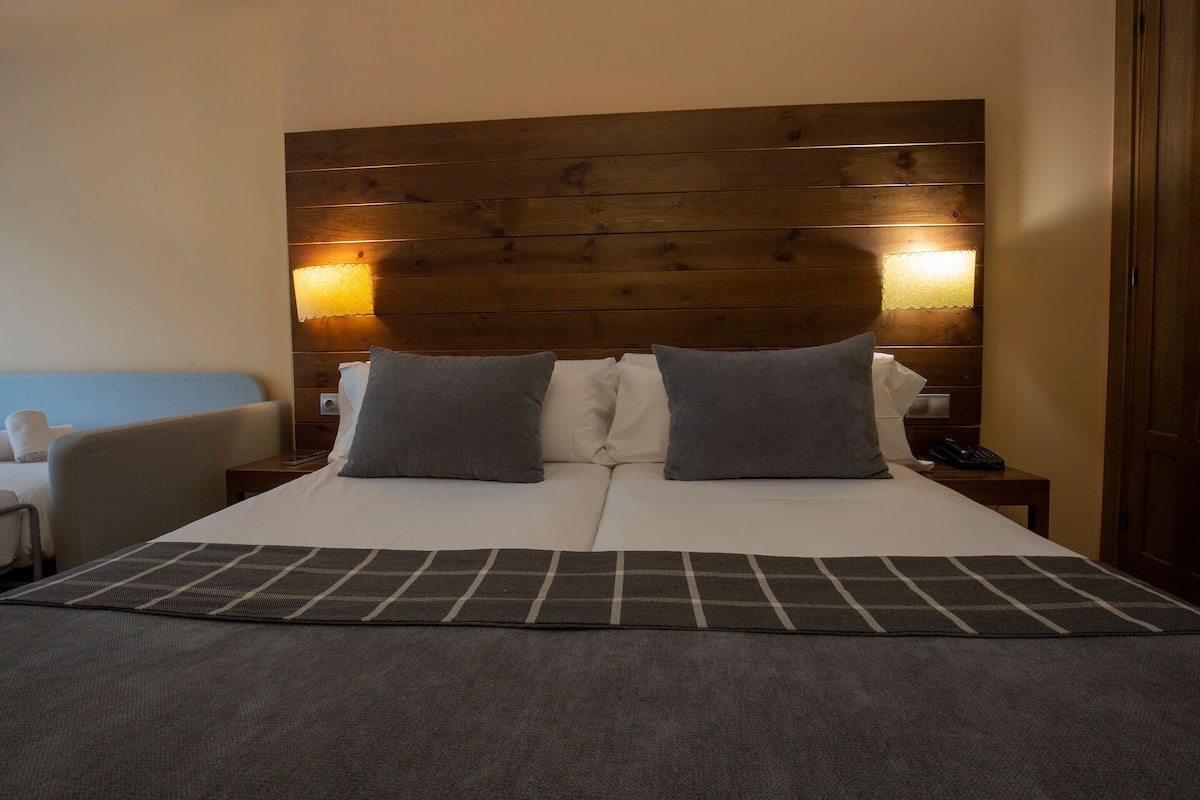 A hotel room with a wooden headboard, two pillows, a bedside table with a lamp, and a bed with a grey blanket.