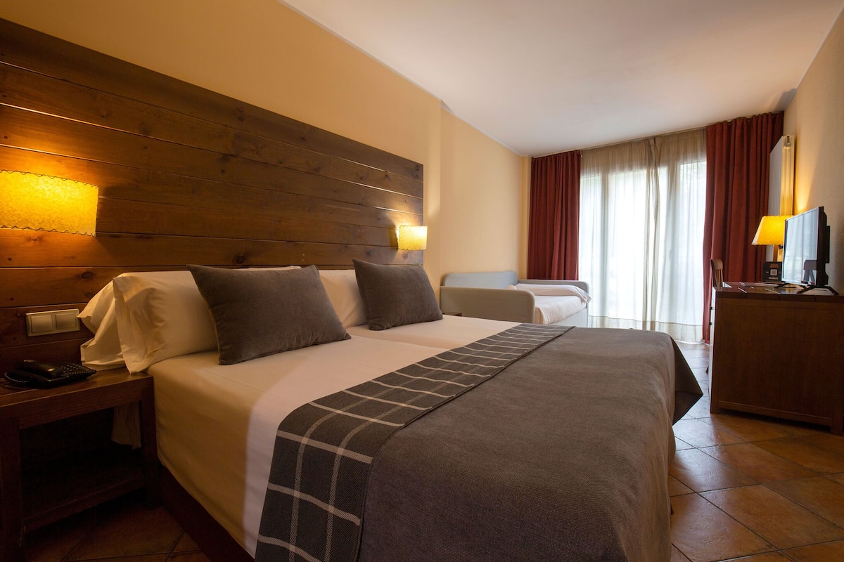 A hotel room with a large bed, a bedside table with a lamp, a desk with a chair, and a window with curtains.