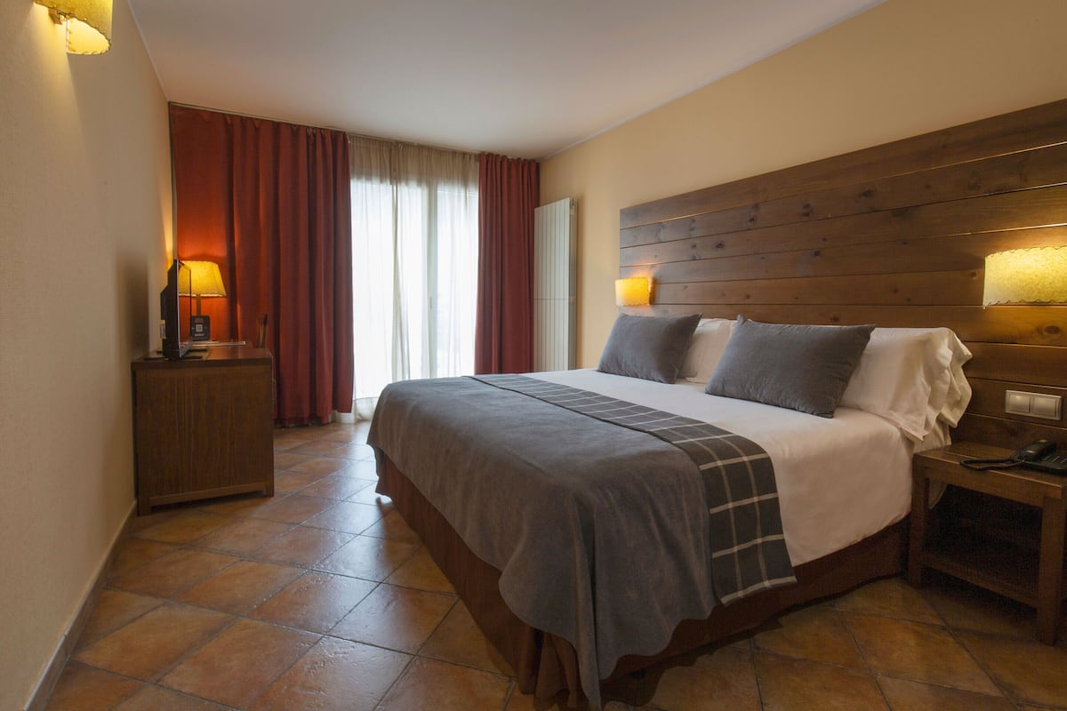 A hotel room with a large bed, wooden headboard, two bedside lamps, a nightstand, and a view of the outdoors through a window with curtains.