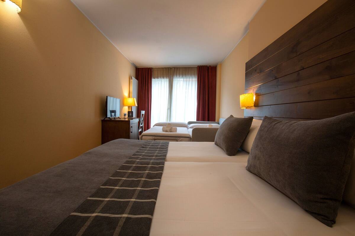 A hotel room with a large bed, a wooden headboard, a bedside table with lamps, and a view of the outdoors through a window with curtains.
