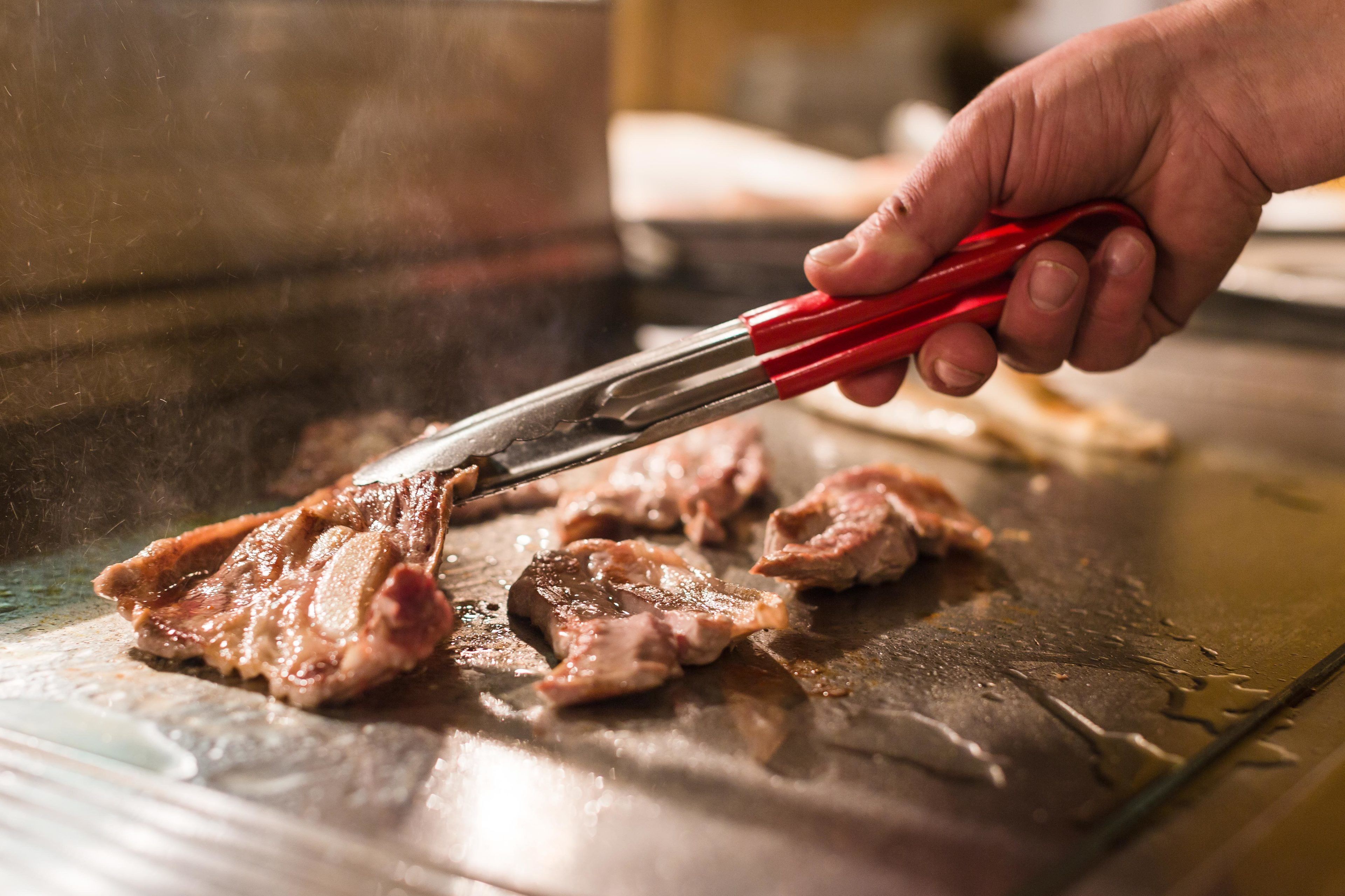 Una mano sosteniendo unas pinzas sobre una parrilla con trozos de carne.